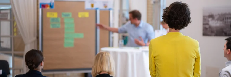 Über die Schultern mehrerer Menschen ist eine Person zu sehen, die an einem Stehtisch steht und auf eine Moderationswand zeigt, an der verschiedene Zettel hängen.