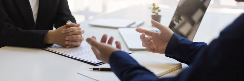 An einem Tisch, auf dem verschieden Zettel und ein Laptop liegen, sitzen sich zwei Personen gegenüber.
