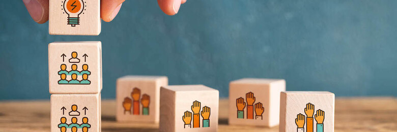 Auf einem Tisch liegen mehrere Holzbauklötze mit Icons, eine Hand baut aus drei Holzbauklötzen einen Turm.