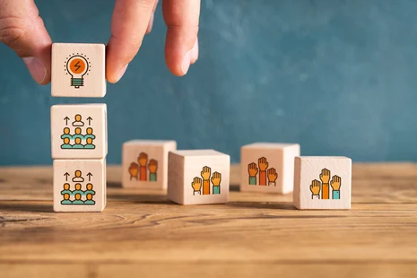 Auf einem Tisch liegen mehrere Holzbauklötze, auf denen verschiedene Icons abgebildet sind; eine Hand baut einen Turm aus drei Holzbauklötzen.