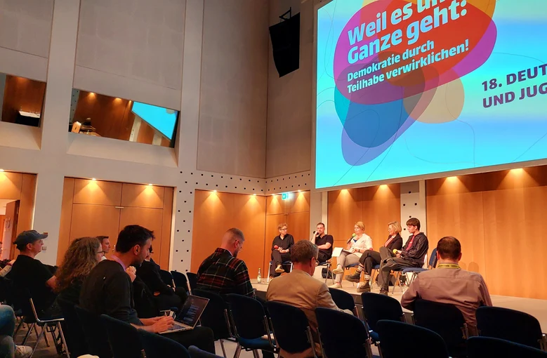 Blick in einem Saal, in dem viele Personen in Stuhlreihen sitzen und zu einem Podium blicken, auf dem fünf Personen sitzen.