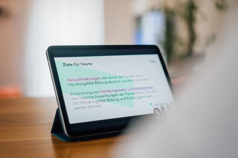 Über die Schulter einer Person ist ein Tablet zu sehen, dass auf einem Tisch steht und auf dem eine Präsentation angezeigt wird.