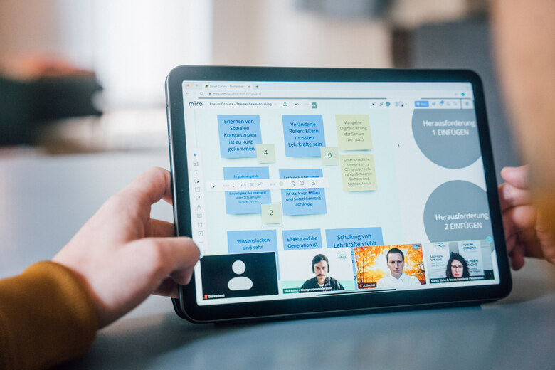 Eine Person hält ein Tablet in der Hand, auf dem sowohl die Teilnehmenden der Videokonferenz als auch eine digitale Pinnwand zu sehen sind.