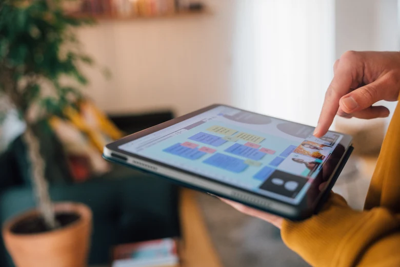 Eine Person hält ein Tablet in der Hand, auf dem sowohl Teilnehmende der Videokonferenz als auch eine digitale Pinnwand zu sehen sind.