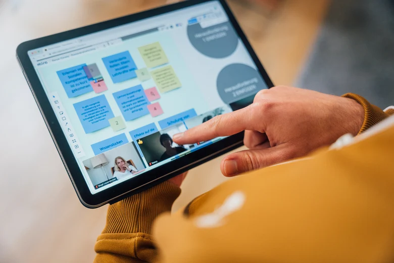 Eine Person hält ein Tablet in der Hand, auf dem die Teilnehmenden der Videokonferenz sowie eine digitale Pinnwand zu sehen sind.