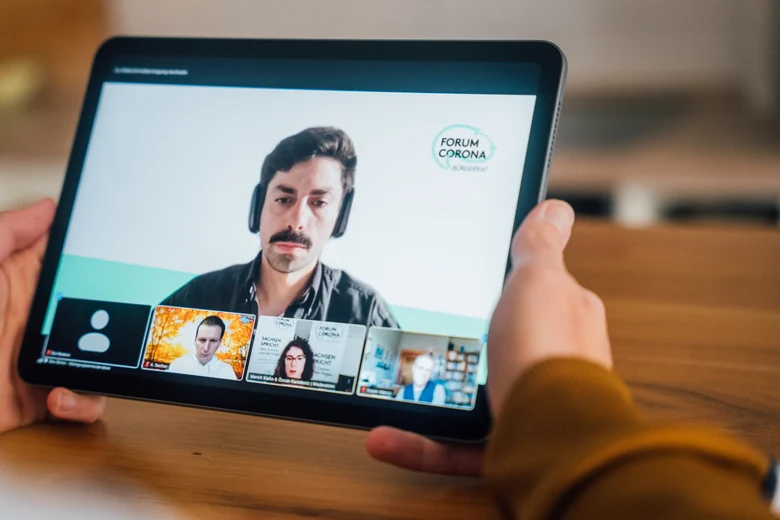 Eine Person hält ein Tablet in der Hand, auf dem eine Videokonferenz geöffnet ist und verschiedene Teilnehmende der Videokonferenz zu sehen sind.