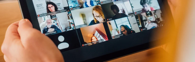 Eine Person hält ein Tablet in den Händen, auf dem verschiedene Teilnehmende einer Videokonferenz zu sehen sind.