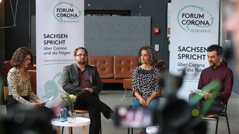 Vier Personen sitzen in einem Halbkreis, hinter ihnen zwei RollUps des Bürgerrats "Forum Corona".