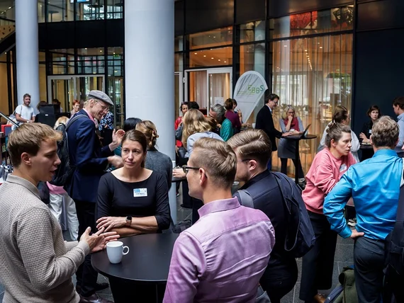 Viele Personen stehen in einem großen Raum und unterhalten sich in kleineren Gruppen an Stehtischen.