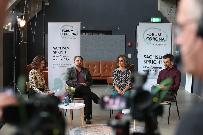 Über die Schulter des Kamerateams sind die vier Podiumsteilnehmenden zu sehen, hinter ihnen stehen zwei RollUps des Forum Corona.