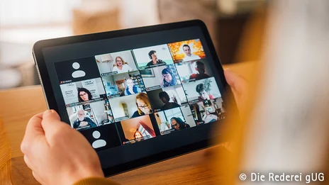 Eine Person hält ein Tablet in der Hand, auf dem in einer Kachelgalerie Videos von verschiedenen Personen in einer Videokonferenz abgebildet sind.