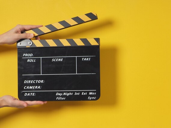 Vor einem gelben Hintergrund wird eine Filmklappe in die Kamera gehalten.