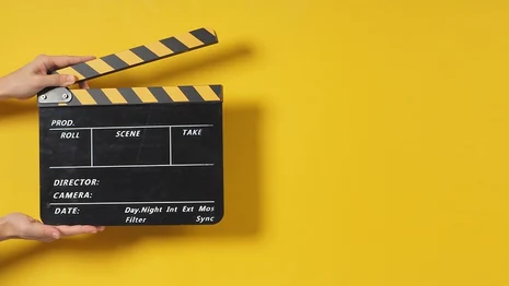 Vor einem gelben Hintergrund hält eine Hand eine schwarze Filmklappe.