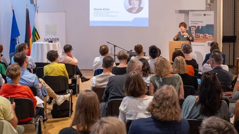 Viele Personen sitzen in Stuhlreihen und blicken zu einem Rednerpult, an dem Gisela Erler spricht.